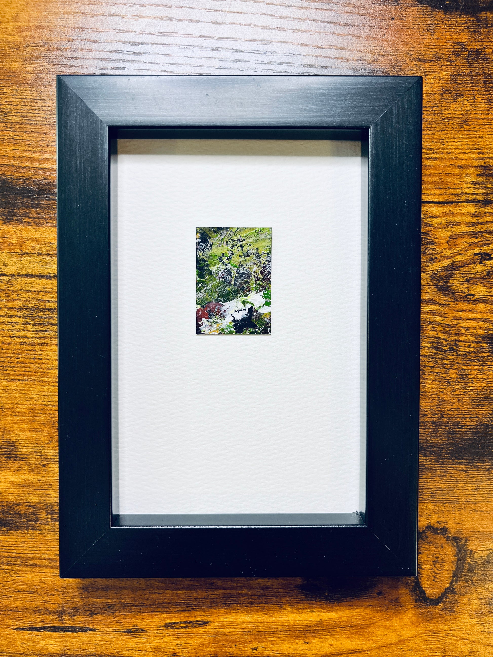 Original artwork by Anet Skillin. This miniature abstract landscape painting features green, yellow ochre, black, white and red colors. Painted on recycled packaging, mounted on 4x6-inch paper and photographed in a black 4x6-inch frame against a wooden table.