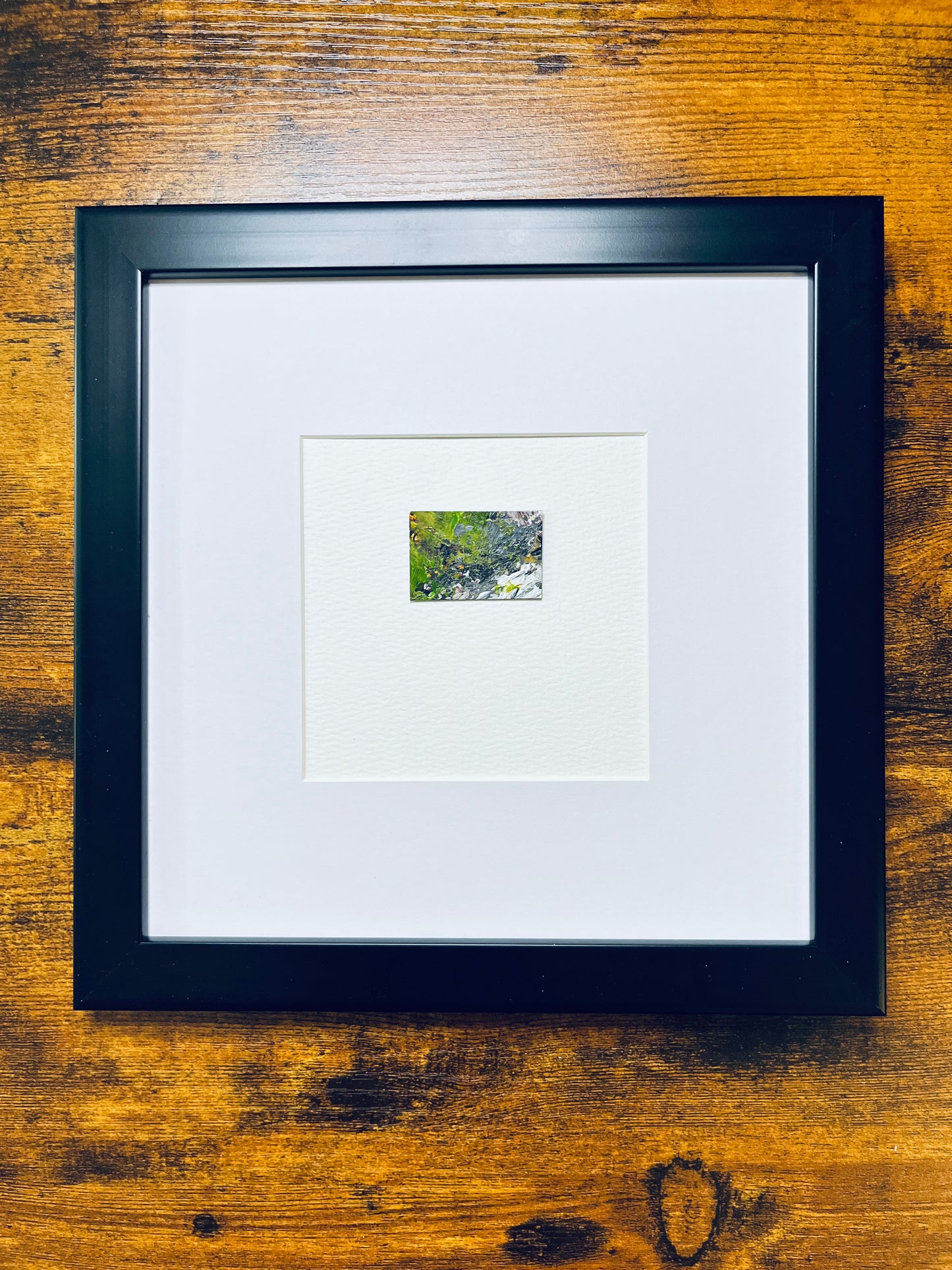 Original artwork by Anet Skillin. This miniature abstract landscape painting features green, yellow ochre, black, white and red colors. Painted on recycled packaging, mounted on 4x6-inch paper and photographed in a black 8x8-inch frame against a wooden table.