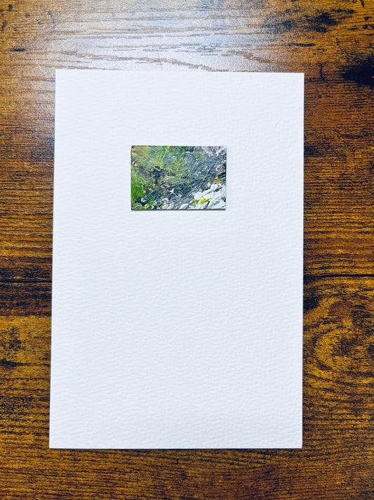 Original artwork by Anet Skillin. This miniature abstract landscape painting features green, yellow ochre, grey, white and pink colors. Painted on recycled packaging, mounted on 4x6-inch paper and photographed against a wooden table.